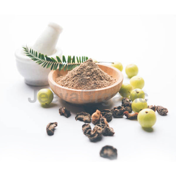 A bowl of herbal powder surrounded by green amla fruits and dried herbs, with a mortar and pestle in the background.