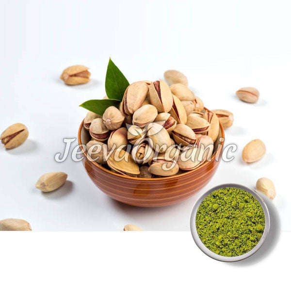 A bowl of pistachios with green leaves, surrounded by scattered nuts and a small bowl of pistachio powder.